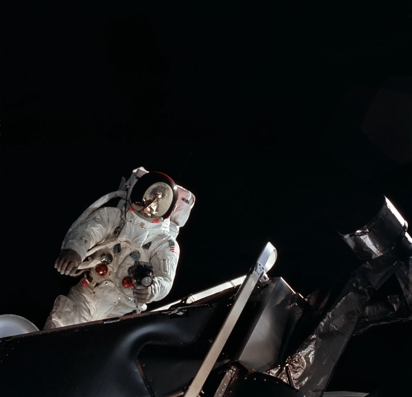An astronaut in a white spacesuit stands in the open hatch of a spacecraft against black space, holding a camera