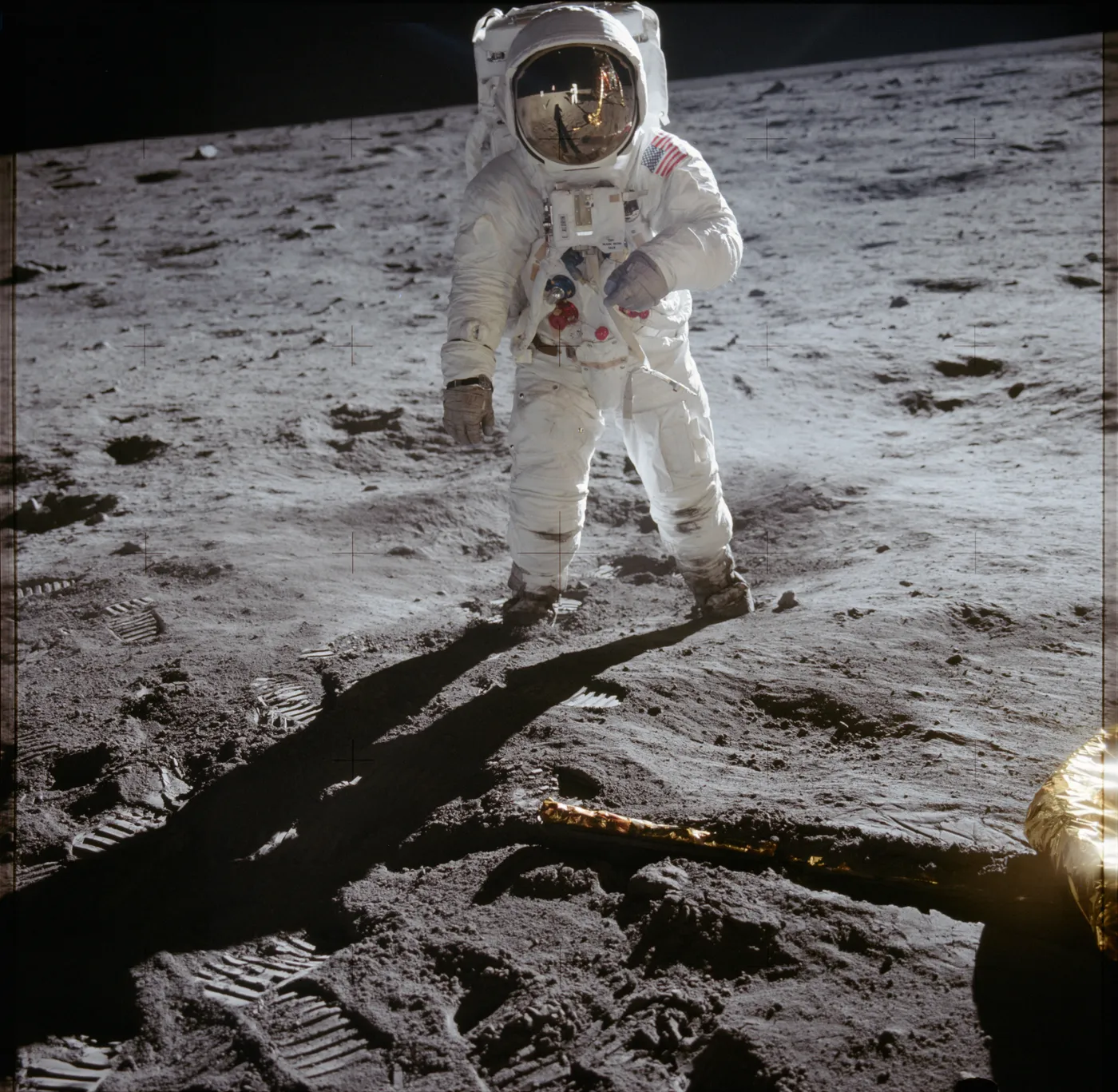 An astronaut stands on the Moon facing the camera; his gold visor reflects the photographer and the lunar lander