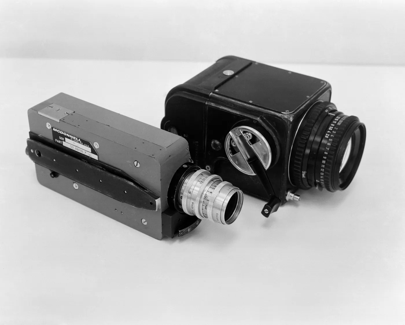 A black Hasselblad camera with a large lens and winding crank, beside a smaller grey 16 mm movie camera, on a white surface